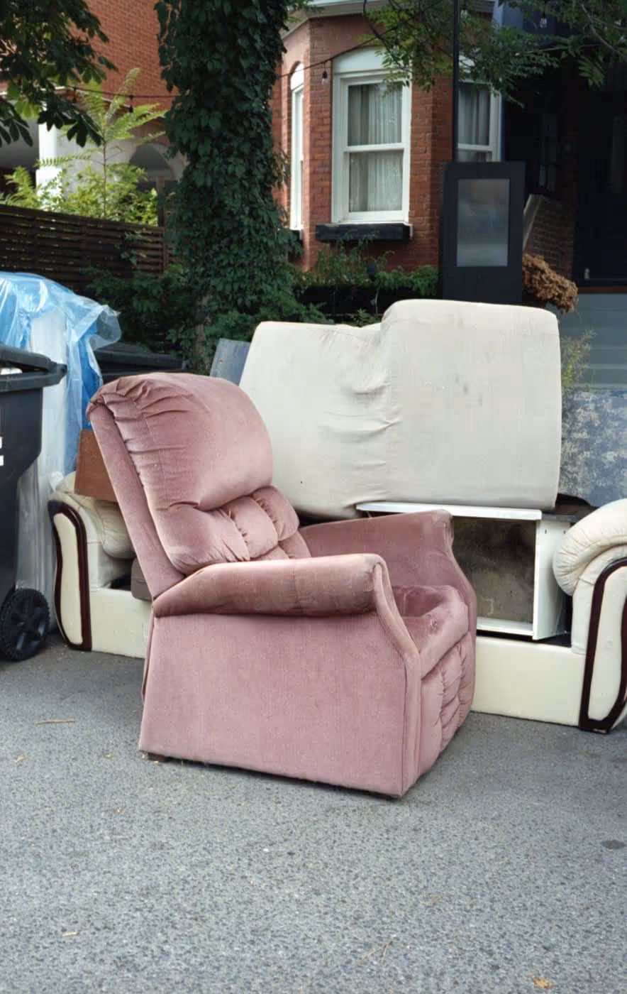 Pink upholstered recliner chair and beige sofa placed on a sidewalk outside a brick house.