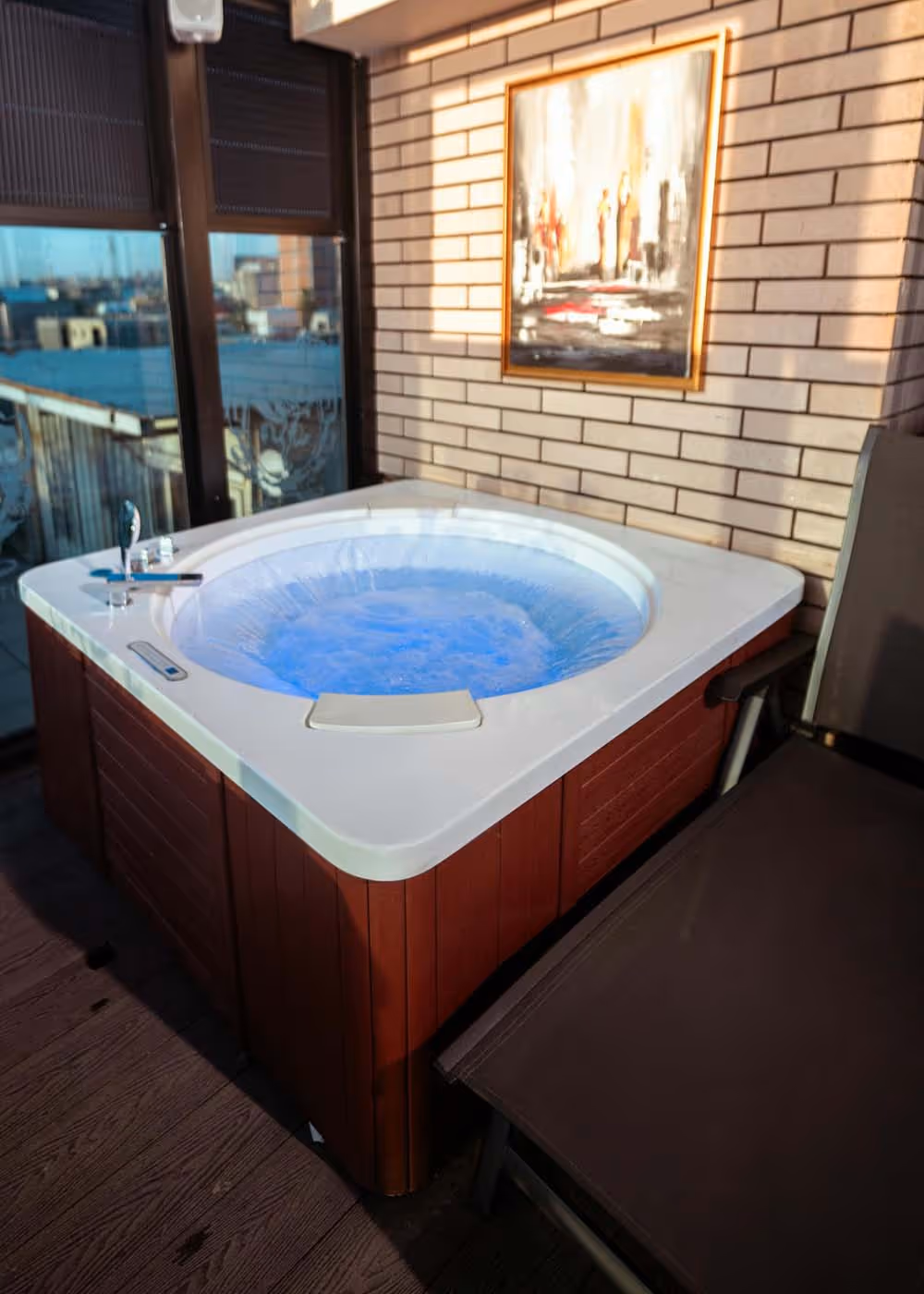 Indoor hot tub with bubbling water and blue lighting next to a lounge chair, set against a brick wall with a framed painting.