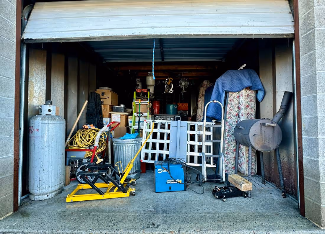 Open garage filled with various equipment including a propane tank, yellow lift, hose, metal trash can, ladder, toolboxes, hand truck, and a small stove.